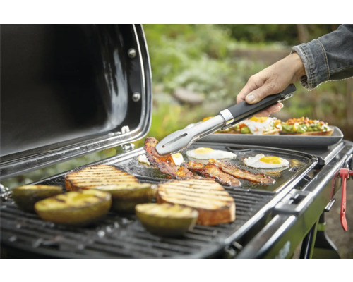 Grill mit Grillgut wie Speck, Eiern, Brot und Avocado, das mit einer Zange gewendet wird.