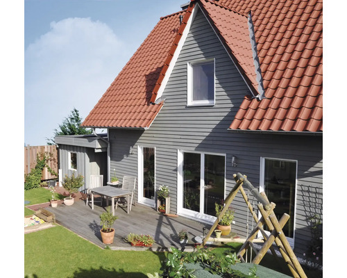 Haus mit grauer horizontaler Fassadenverkleidung, rotem Ziegeldach und einer Holzterrasse mit Tisch, Stühlen und Topfpflanzen neben einer Rasenfläche.