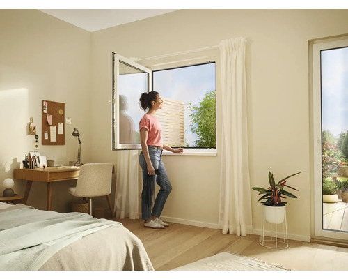 Helles Zimmer mit geöffnetem Fenster, Schreibtisch, Bett und einer Frau, die zum Fenster hinausschaut