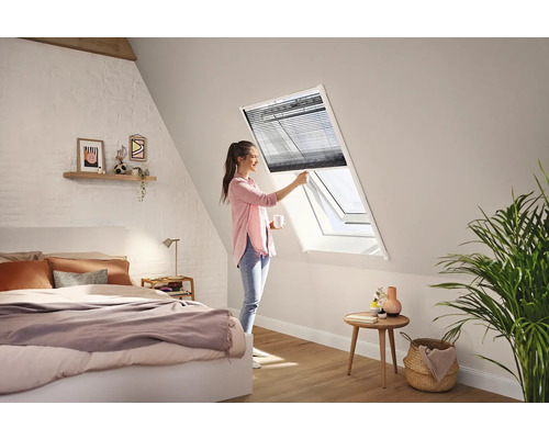 Eine Frau bedient ein dunkles Plissee an einem weißen Dachfenster in einem hellen, modernen Schlafzimmer mit Dachschräge.