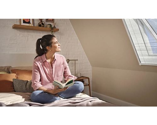 Frau liest auf einem Bett in einem hellen Dachzimmer mit Dachfenster-Jalousie, weißer Ziegelwand und Holzregal.