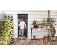 Ein Mann geht durch einen magnetischen Insektenschutzvorhang an einer Terrassentür auf eine Holzterrasse mit verschiedenen Topfpflanzen.