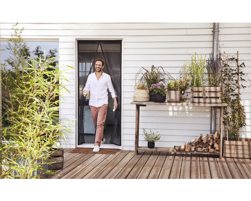 Ein Mann geht durch einen magnetischen Insektenschutzvorhang an einer Terrassentür auf eine helle Holzterrasse mit vielen Topfpflanzen.