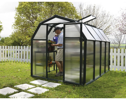 Kleines Gewächshaus in Scheunenform mit schwarzem Rahmen und Polycarbonat-Paneelen im Garten. Ein Mann gießt im Inneren Pflanzen.