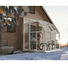 Weißes Anlehn-Gewächshaus mit Glaswänden an einem Haus in einer Schneelandschaft. Eine Person liest im hellen, eingerichteten Innenraum.