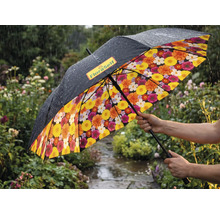 Regenschirm in einem regnerischen Garten mit schwarzer Außenseite, FloraSelf Logo und buntem Blumenmuster auf der Innenseite.