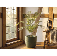 Künstliche Palme in einem grünen strukturierten Topf in einem hellen, rustikalen Raum mit Holzbalken, einem Fenster und einem Hocker mit Laterne.