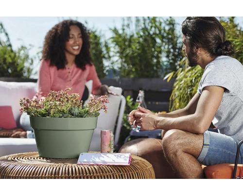Zwei Personen sitzen auf einer Terrasse an einem runden Rattantisch mit einem hellgrünen Blumentopf mit Sukkulenten vor grüner Gartenkulisse.