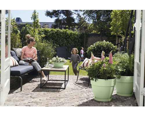 Moderne Terrasse mit Lounge-Sofa, Holztisch und hellgrünen Pflanztöpfen. Frau auf dem Sofa, Kind auf dem Rasen im Hintergrund.