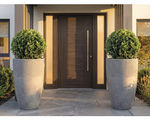Moderne Haustür in Holzoptik mit langem Metallgriff, flankiert von zwei matten Seitenteilen und großen grauen Pflanzkübeln mit Formgehölz.