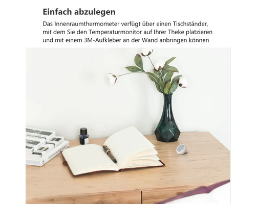 Kleines rundes Innenthermometer auf einem Holzschreibtisch neben einem Notizbuch und einer Vase mit Blumen.