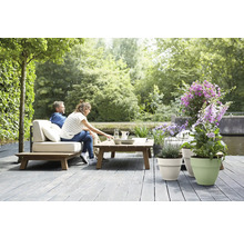 Helles, geräumiges Lounge-Set aus Holz mit hellen Polstern auf einer grauen Terrasse am Wasser, dekoriert mit Pflanzgefäßen und Blumen.