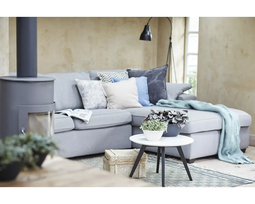 Modernes Wohnzimmer mit grauem Ecksofa, Zierkissen, hellblauer Wohndecke, weißem Beistelltisch mit Pflanzen und einem grauen Kaminofen.