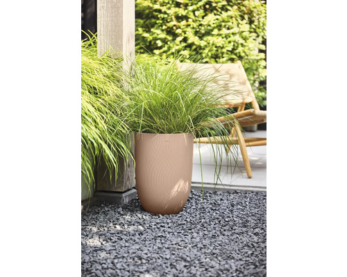 Hoher runder Pflanzkübel mit vertikaler Wellenstruktur und Ziergras auf einer Kiesfläche im Garten neben einem Holzpfosten und einem Gartenstuhl.