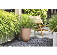 Brauner runder Pflanzkübel mit geriffelter Struktur und grünem Ziergras auf einer Terrasse mit dunklem Kies neben einem Holzstuhl.