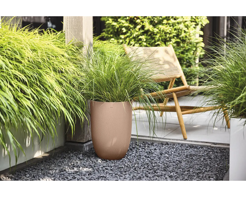 Brauner runder Pflanzkübel mit geriffelter Struktur und grünem Ziergras auf einer Terrasse mit dunklem Kies neben einem Holzstuhl.