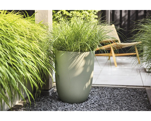 Moderner, hoher Pflanzkübel mit vertikaler Wellenstruktur und Ziergras auf einer hellen, organisierten Terrasse mit Kies und Bodenplatten.