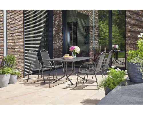 Vier Gartenstühle aus grauem Streckmetall stehen um einen passenden Tisch auf einer hell gepflasterten Terrasse vor einer Klinkerwand.