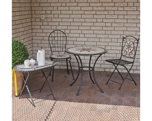 Bistro-Set mit rundem Mosaiktisch und zwei Metallstühlen auf einer Terrasse vor einer Ziegelwand. Ein passender ovaler Beistelltisch steht daneben.