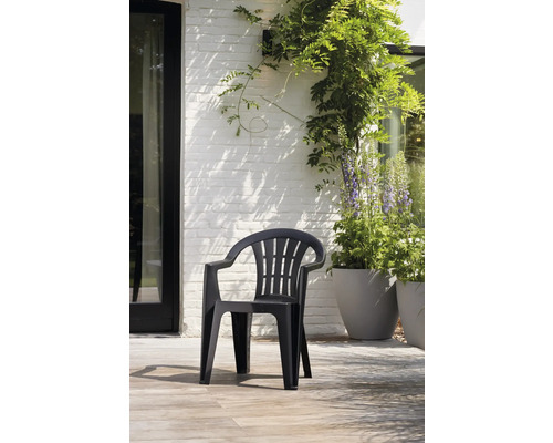 Stapelbarer Gartenstuhl aus schwarzem Kunststoff mit Armlehnen auf einer hellen Terrasse vor einer weißen Ziegelwand mit Pflanzen.