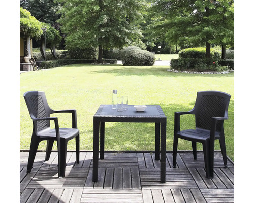 Zwei anthrazitfarbene Gartenstühle und ein quadratischer Tisch in Rattanoptik auf einer Holzterrasse vor einem grünen Garten mit Rasen und Bäumen.