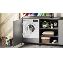 Einbau-Waschmaschine mit offener Tür in grauem Schrank mit Regalen, Handtüchern, Pflanze und Wäschekorb im hellen Hauswirtschaftsraum.