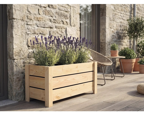 Rechteckiger Pflanzkasten aus Holzlamellen mit blühendem Lavendel auf einer Holzterrasse vor einer Natursteinwand.