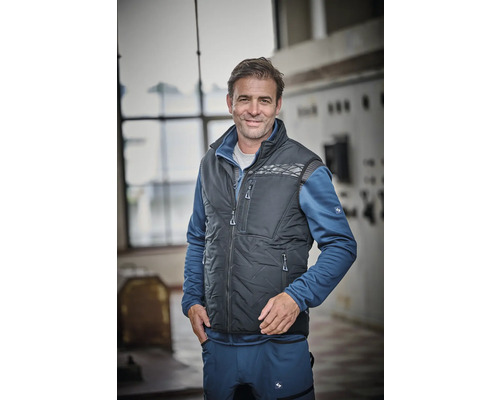 Mann trägt eine dunkelgraue Steppweste über einer blauen Fleecejacke und einer blauen Arbeitshose.