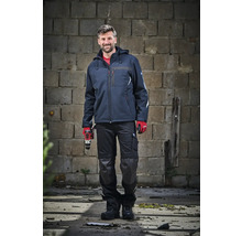 Ein Mann in dunkelblauer Arbeitsjacke und schwarzer Arbeitshose mit Kniepolstern hält einen Akkuschrauber vor einer Steinwand.