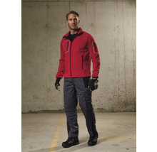 Mann in roter Arbeitsjacke, grauer Arbeitshose, schwarzen Arbeitshandschuhen und schwarzen Sicherheitsschuhen vor einer Betonwand.
