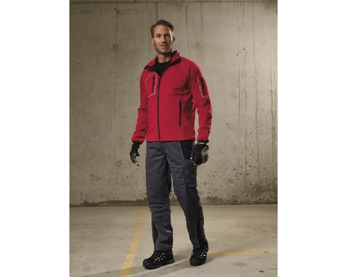 Mann in roter Arbeitsjacke, grauer Arbeitshose, schwarzen Arbeitshandschuhen und schwarzen Sicherheitsschuhen vor einer Betonwand.