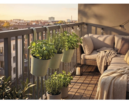 Balkon mit Kräutertöpfen am Geländer, Loungesofa mit Kissen und Decke, Holzfliesen, Laterne und Lichterkette vor einem Stadtpanorama.