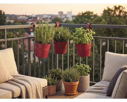 Rote Balkonhängetöpfe aus Metall mit Kräutern an einem Balkongeländer, umgeben von Loungemöbeln und weiteren Pflanztöpfen auf einem Holzboden.