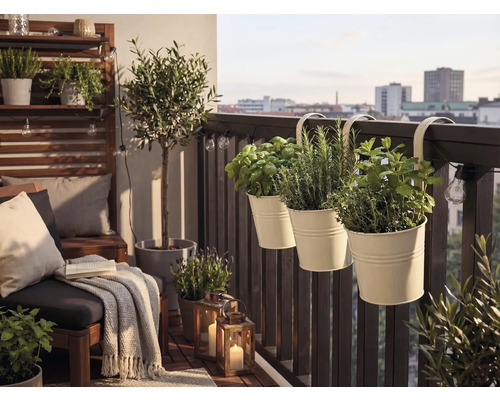 Hell gestalteter Balkon mit hängenden Kräutertöpfen am Geländer, Loungemöbeln, einem kleinen Olivenbaum und dekorativen Laternen.