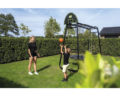 Kind wirft Basketball in Korb an schwarzem EXIT Toys Klettergerüst mit Nestschaukel und Schaukel im Garten.