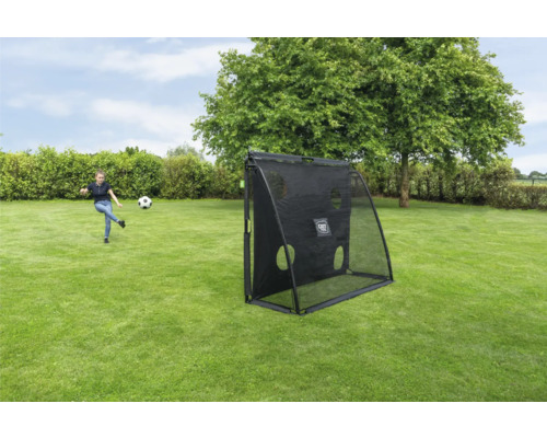 Mädchen spielt Fußball mit einem schwarzen EXIT Toys Rebounder mit Zielwand auf einer grünen Wiese im Garten.