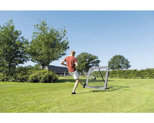 Person schießt Fußball gegen ein Rebounder-Netz in einem weitläufigen Garten mit Bäumen unter freiem Himmel.