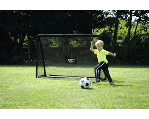 Kind spielt mit Fußball vor schwarzem EXIT TOYS Fußballtor auf Rasenfläche mit Bäumen im Hintergrund.
