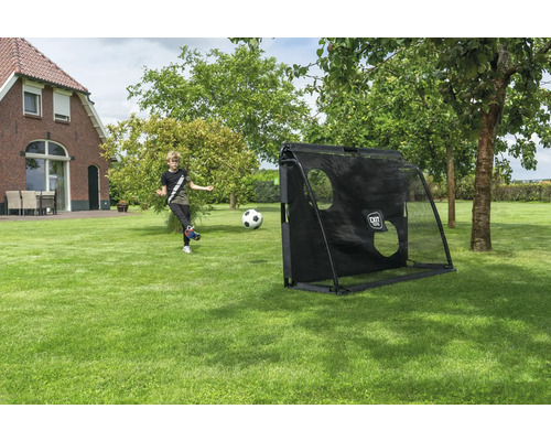 Ein Junge schießt einen Fußball auf ein schwarzes Fußballtor mit EXIT TOYS Logo und Torwandplane in einem großen Garten mit Rasen und Bäumen.