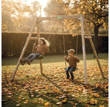 Holz-Doppelschaukel mit zwei schaukelnden Kindern in einem hellen Garten mit herbstlichem Laub.