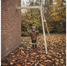 Kind auf einer wandmontierten EXIT Toys Einzelschaukel aus Holz in einem Garten mit Herbstlaub vor einer Ziegelwand.