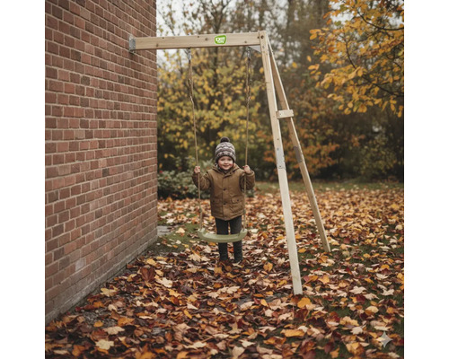 Kind auf einer wandmontierten EXIT Toys Einzelschaukel aus Holz in einem Garten mit Herbstlaub vor einer Ziegelwand.