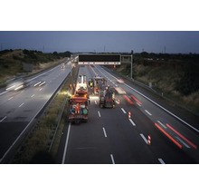 Straßenbauarbeiten auf einer mehrspurigen Autobahn bei Nacht mit Baustellenfahrzeugen, Arbeitern in Warnschutzkleidung und Leitkegeln.