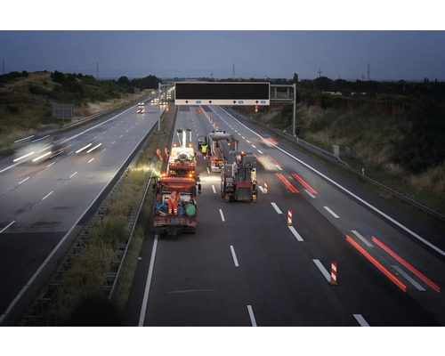 Straßenbauarbeiten auf einer mehrspurigen Autobahn bei Nacht mit Baustellenfahrzeugen, Arbeitern in Warnschutzkleidung und Leitkegeln.