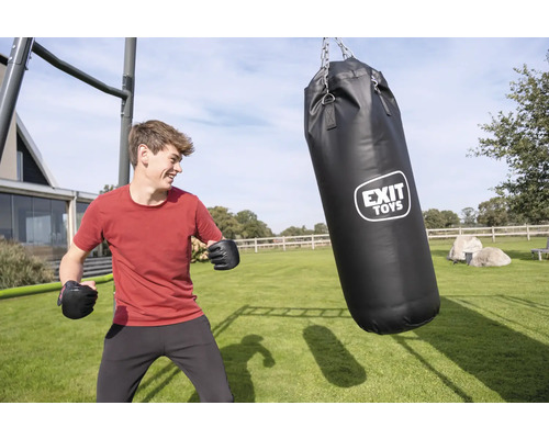 Ein junger Mann mit Boxhandschuhen im Garten neben einem schwarzen EXIT TOYS Boxsack, der an einem Metallrahmen auf einem hellen Rasen hängt.