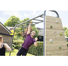 Ein Mann trainiert an einem grauen Fitnessgerüst aus Metall mit Hangelstangen und Holzkletterwand in einem hellen Garten.