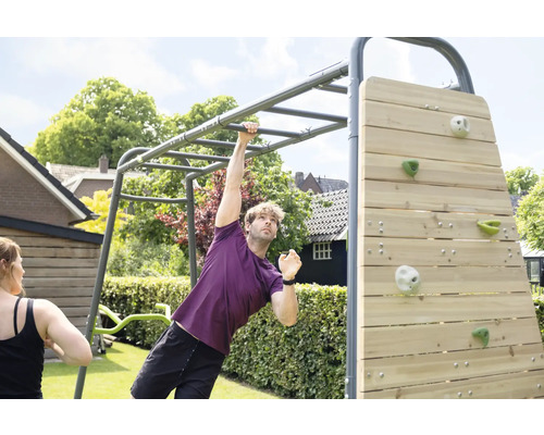 Ein Mann trainiert an einem grauen Fitnessgerüst aus Metall mit Hangelstangen und Holzkletterwand in einem hellen Garten.