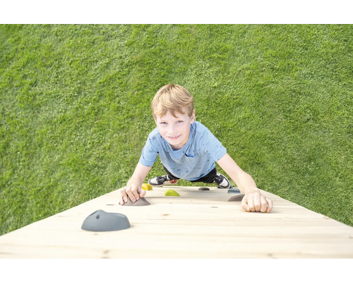 Ein Kind klettert an einer Kletterwand aus Holz mit bunten Klettersteinen auf einer Rasenfläche.