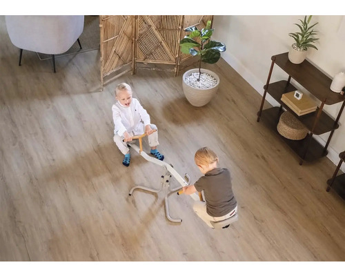 Zwei Kinder spielen auf einer Wippe in einem hellen Zimmer mit einem Bodenbelag in Holzoptik.