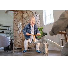 Ein Kind sitzt auf einer Metallwippe in einem hellen, modern eingerichteten Raum mit einem Rattan-Paravent und Zimmerpflanzen.